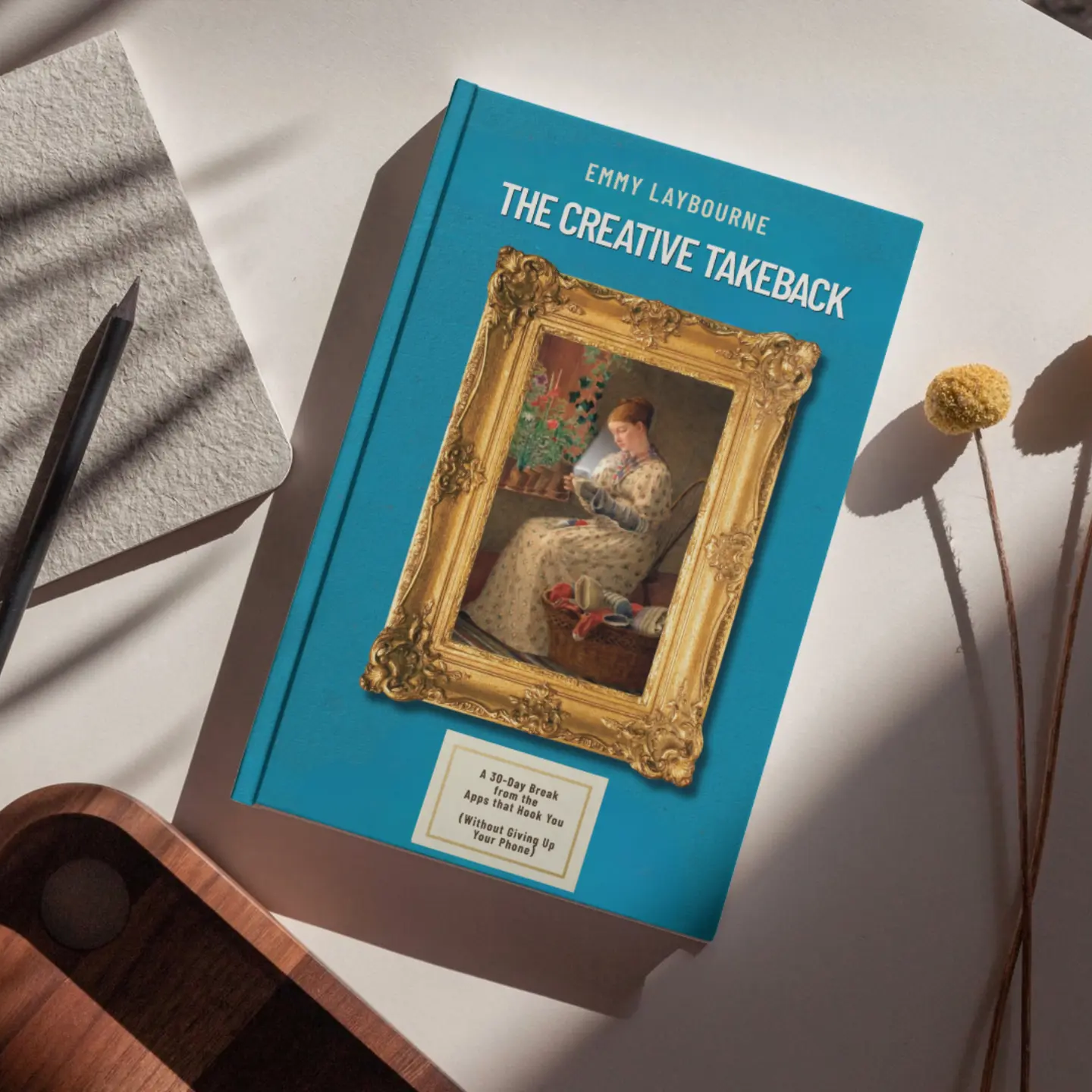 The Creative Takeback book — a teal hardcover with an ornate gold frame holding a painting of a woman reading, resting on a table beside a notebook, pencil and dried flowers